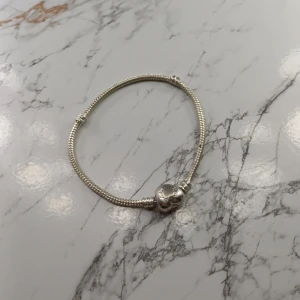 Pandora armband med hjärtformad lås - Snyggt armband från Pandora i silverfärgat material med ett hjärtformat lås där Pandora-loggan är ingraverad. Armbandet har en klassisk, rundad form och passar perfekt för att kombinera med olika berlocker. Stilren och tidlös design.