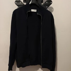 Mörkblå kofta från Selected Homme - Snygg mörkblå kofta från Selected Homme med ribbad krage och hel dragkedja framtill. Tröjan är stickad i mjukt material och har långa ärmar. Perfekt för dig som gillar stilrena och klassiska plagg med en modern touch.