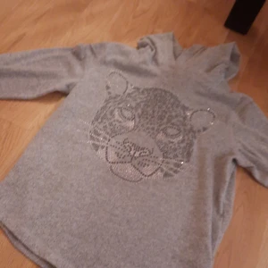 Grå hoodie med leopardmotiv - Säljer en grå hoodie med huva och lång ärm. Framsidan har ett stort leopardmotiv gjort av glittriga stenar som verkligen sticker ut. Perfekt för dig som gillar djurmönster och vill ha en chill men snygg look.