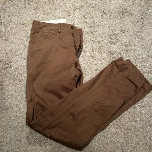 Bruna chinos från Uniqlo, storlek 31/34 - Snygga bruna chinos från Uniqlo i storlek 31/34. Byxorna har klassisk rak passform, midwaist och är tillverkade i 100% bomull. Perfekta för dig som gillar stilrena och bekväma byxor med enkel design och diskreta fickor.