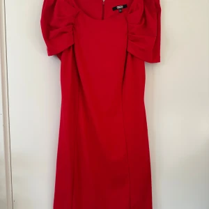 Röd klänning med puffärm från ASOS - Säljer en röd klänning från ASOS med snygga puffärmar och draperade detaljer vid axlarna. Klänningen har rund halsringning och är tillverkad i ett mjukt tyg som ger en skön känsla. Perfekt för dig som vill sticka ut med en färgstark look.