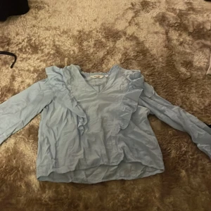 Ljusblå blus med volangdetaljer - Supersöt ljusblå blus med långa ärmar och v-ringning. Blusen har snygga volangdetaljer framtill och längs axlarna som ger en trendig look. Perfekt att styla med jeans eller kjol för en fräsch outfit.