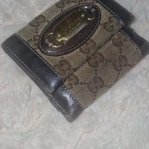 Plånbok  - Snygg brun plånbok från med klassiskt GG-mönster och detaljer i mörkbrunt skinn. Plånboken har flera kortfack, myntfack och en metallskylt framtill. Perfekt för dig som vill ha en stilren och lyxig accessoar. Finns synliga tecken på användning men inget som är trasigt.