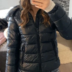 Svart pufferjacka med dragkedja - Snygg svart pufferjacka med quiltad design och dragkedja framtill. Jackan har hög krage och två fickor med dragkedja på sidorna. Perfekt för kyliga dagar och ger en cool, sportig vibe.