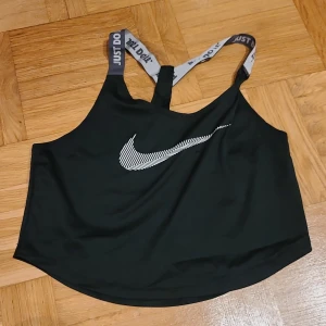 Svart Nike Dri-Fit linnetopp XS - Svart linnetopp från Nike i Dri-Fit material med grå axelband där det står 'Just Do It'. Framsidan har en vit Nike-logga med coolt mönster. Perfekt för träning och sportiga outfits, sitter skönt med racerback och ärmlös design.