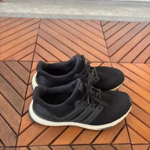 Svarta Adidas Ultraboost sneakers - Svarta Adidas Ultraboost sneakers med stickad ovandel och klassiska tre ränder på sidan. Skorna har snörning och en vit, dämpad Boost-sula som ger grym komfort. Perfekta för dig som gillar sportig stil och vill ha riktigt sköna skor till vardagen.