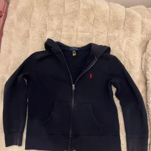 Mörkblå hoodie från Polo Ralph Lauren - Klassisk mörklblå hoodie från Polo Ralph Lauren med dragkedja framtill och huva. Tröjan har en röd broderad logga på bröstet och två fickor framtill. Perfekt för en chill och stilren look.