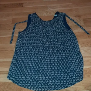 Mönstrad ärmlös blus från H&M - Mörkblå ärmlös blus från H&M med vitt grafiskt mönster och knytband i midjan. Blusen har rund halsringning med dekorativ spetskant och är tillverkad i ett lätt och luftigt material, perfekt för varma dagar.