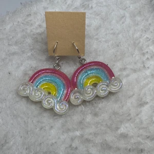 Glittriga regnbågsörhängen - Färgglada örhängen i form av regnbågar med glittriga moln. Örhängena har rosa, blå och gul färg med skimrande detaljer som gör dem extra iögonfallande. Perfekt för dig som vill sticka ut och sprida good vibes.