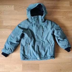 Grön vinterjacka Polarn O. Pyret  - Snygg grön vinterjacka från Polarn O. Pyret med huva, dragkedja och flera fickor. Jackan har svarta muddar vid ärmsluten och reflexdetaljer på ärm och huva. Perfekt för kalla dagar och håller värmen bra. Storlek 116. 5-6 år.