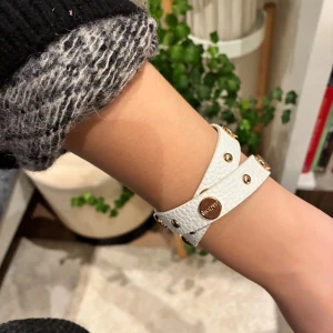 Vitt faux-läder armband IsaDora - Snyggt vitt armband från IsaDora med guldiga metalldetaljer. Armbandet är dubbelt och har en cool, modern look som passar perfekt till din outfit. Aldrig använt!