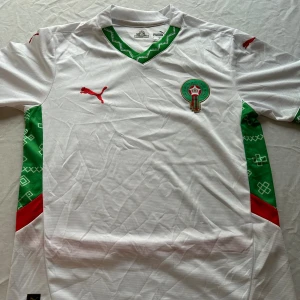 Marocko fotbollströja Puma vit/grön - Marockos officiella fotbollströja  Puma i vitt med gröna och röda detaljer. Tröjan har korta ärmar, grönt mönster längs sidorna och halsen, samt broderad landslagsemblem och Puma-logga. Tillverkad i lätt och ventilerande polyester.