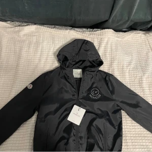 Svart vindjacka från Moncler med huva - Svart vindjacka från Moncler med huva och dragkedja framtill. Jackan har Moncler-logga på bröstet och ärmen, samt två fickor med dragkedja. Tillverkad i glansig polyester som ger en clean och sportig look.