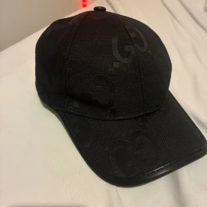 Svart keps från Gucci - Snygg svart keps från Gucci med diskret GG-monogram över hela kepsen. Kepsen är tillverkad i bomull och polyester med detaljer i skinn, justerbar rem baktill och klassisk böjd skärm. Perfekt för dig som vill ha en lyxig och stilren accessoar.