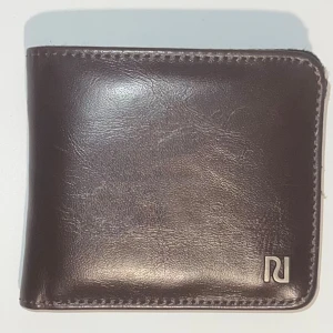 Brun plånbok från River Island - Snygg brun plånbok från River Island i konstläder med stilren design. Plånboken har flera kortfack, myntfack med dragkedja och tryckknapp. Perfekt för dig som vill ha en klassisk och praktisk accessoar med diskret metallogo i hörnet.