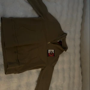 Tommy HilfigerHoodie ZIP MOCK - Snygg beige hoodie från Tommy Hilfiger med half zip och broderad logga på bröstet. Tröjan har långärmade ärmar, stor ficka framtill och Hilfiger-tryck på ena ärmen. Tillverkad i ekologisk bomull för extra skön känsla.