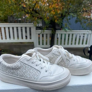 Vita sneakers från Tommy Hilfiger - Snygga vita sneakers från Tommy Hilfiger med diskret mönstrade sidor och klassisk snörning. Skorna har rund tå, platt sula och detaljer i både skinn och textil. Perfekta för dig som gillar stilrena och fräscha sneakers med en twist.