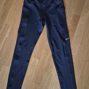 Svarta Nike leggings med fickor, S - Snygga svarta leggings från Nike i storlek S, med höga fickor på sidorna och klassisk Nike-logga på benet. Tillverkade i stretchigt material med 82% polyester och 18% elastan för skön passform. Perfekta för träning eller chill.
