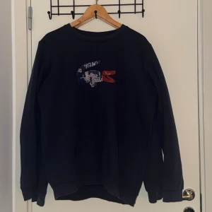 Svart sweatshirt från Lacoste - Svart sweatshirt från Lacoste med broderad krokodil-logga i blått, vitt och rött på bröstet. Tröjan har rund hals, långa ärmar och ribbade muddar vid ärmslut och nederkant. Perfekt för en chill och stilren look.