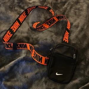 Nike svart axelväska - Snygg svart axelväska från Nike med bredt orange/ svart axelband där det står NIKE i svart eller orange beroende på sida. Väskan har flera fack, dragkedjor med detaljer och mesh på baksidan för extra komfort. Inköpt på Zalando