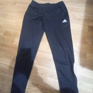 Svarta Adidas mjukisbyxor - Svarta mjukisbyxor från Adidas med klassisk logga på låret. Byxorna har elastisk midja och är tillverkade i mjukt syntetmaterial, perfekta för chill eller träning. Raka ben och enkel design gör dem lätta att matcha med det mesta.