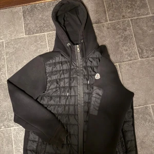 Svart pufferjacka från Moncler - Svart pufferjacka från Moncler med huva och snörning. Jackan har quiltad front, mjuka ärmar och dragkedja hela vägen. Moncler-logga på vänster ärm och praktiska fickor med dragkedja. Perfekt för dig som gillar stilren och sportig look. Drag kedjan är lite trasig men ändå bra 
