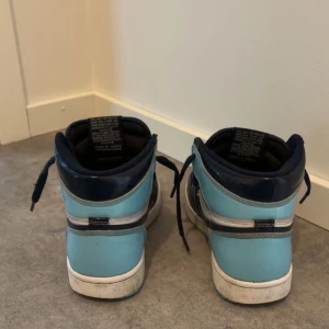 Nike Air Jordan 1 High blå/svart - Nike Air Jordan 1 High sneakers i blått och svart med vit sula. Klassisk high-top siluett med perforerad tå, snörning och rund tå. Skorna har en cool färgkombo och är tillverkade i skinn med syntetdetaljer. Perfekta för dig som gillar streetwear och sneakers med attityd.