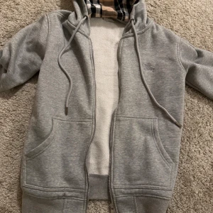 Grå Burberry hoodie med rutig huva - Säljer en grå hoodie från Burberry i storlek S. Tröjan har dragkedja, snörning och huva med klassiskt Burberry-rutigt foder i beige, svart och rött. Broderad logga på bröstet och två fickor framtill. Perfekt för dig som gillar stilrena och lyxiga detaljer. OBS zippern funkar inte men går att fixa