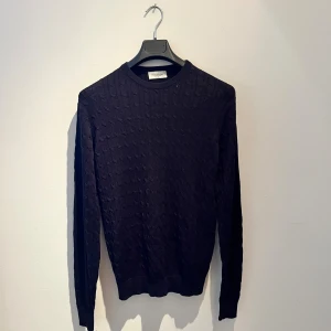 Mörkblå stickad tröja Bruun & Stengade - Mörkblå stickad tröja från Bruun & Stengade i storlek M. Tröjan har ett diskret kabelstickat mönster och rund hals. Mjukt material och långärmad passform som sitter snyggt på kroppen. Perfekt för dig som gillar stilrena och klassiska plagg.
