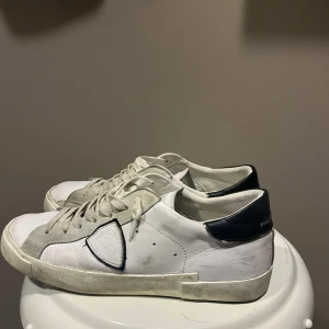 Philippe Model - Snygga vita sneakers från Philippe Model med svarta detaljer på hälen och grå mockadetaljer längs snörningen. Skorna har klassisk låg profil, rund tå och platt sula. Tillverkade i skinn och mocka med tydlig logga på sidan och bak på hälen.