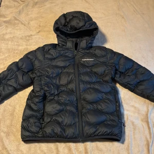 Svart dunjacka Peak Performance 170 - Svart dunjacka från Peak Performance i storlek 170. Jackan har huva, dragkedja framtill och två sidofickor med dragkedja. Quiltad design med snygg struktur och klassisk logga på bröstet. Perfekt för kalla dagar och riktigt skön att bära. Ända defekten är ett litet håll längst ner på armen