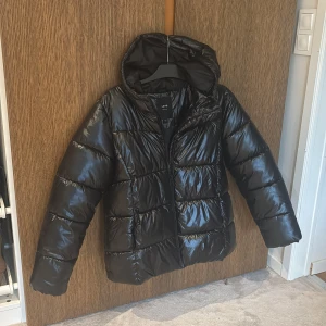 Svart pufferjacka med huva från kids brandstore, märke (LMTD) - Säljer en svart, glansig pufferjacka från LMTF med huva och dragkedja. Jackan är quiltad och har en normal puffjacka passform som ger en cool och avslappnad vibe. Perfekt för kalla dagar när du vill vara både varm och snygg. Obs slutsåld på nätet/butik ny pris 799kr
