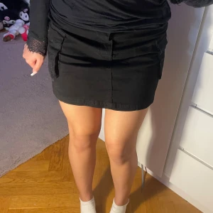 Mini cargoskirt - Snygg svart cargokjol med fickor och knappar – perfekt mix av street och classy. Tjockare bomullstyg som håller formen och sitter fint på kroppen. Storlek: M passar även S Helt ny med tag kvar ✨  🖤 Perfekt till hoodie, croptop eller boots för en cool vardagslook!
