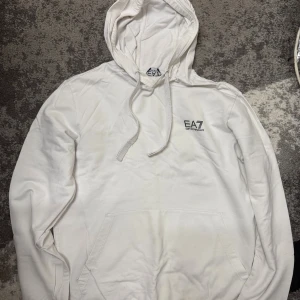 Vit hoodie från EA7 Emporio Armani - Säljer en fräsch vit hoodie från EA7 Emporio Armani med diskret logga på bröstet. Tröjan har huva med snörning och en stor magficka. Perfekt för chill dagar och enkel att matcha med jeans eller joggers.