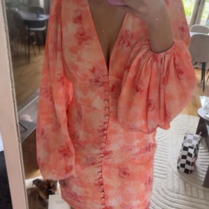 Adoore klänning - Superfin klänning i ljusa rosa och orange toner med blommigt mönster. Klänningen har djup v-ringning, puffiga långärmar och knappar längs hela framsidan. Materialet är lätt och flowy, perfekt för sommarens alla tillfällen.