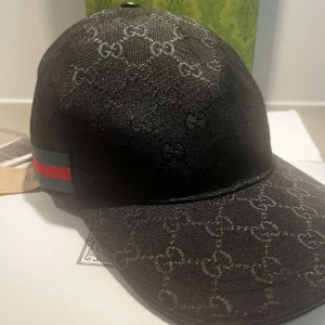 Svart keps från Gucci med GG-mönster - Säljer en svart keps från Gucci med klassiskt GG-monogram över hela kepsen. Sidan har en ikonisk grön och röd rand. Kepsen är tillverkad i bomull och polyester, perfekt för dig som vill ha en stilren och lyxig accessoar.