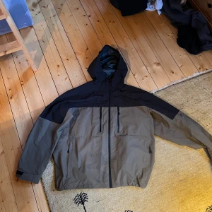 Svart och beige vindjacka med huva H&M - Säljer en stilren vindjacka i svart och beige med huva och hel dragkedja framtill. Jackan har två vertikala bröstfickor med dragkedja och stora sidofickor. Perfekt för dig som gillar funktionell och snygg streetwear.