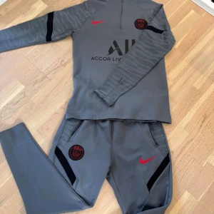 PSG Tracksuit - Snygg PSG långärmad träningströja från Nike i mörkgrått med svarta och röda detaljer. Tröjan har halv dragkedja, svart Paris Saint-Germain-logga med röd brodyr, och subtilt zebramönster på ärmarna. Pris kan diskuteras 