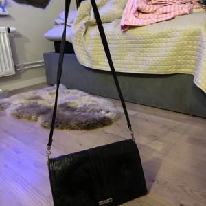 Svart axelväska i skinn med dragkedja - Snygg svart axelväska i skinn med lång rem och stilren design. Väskan har ett praktiskt innerfack med dragkedja och ett lock som stängs framtill. Perfekt storlek för att få plats med det viktigaste och passar till många olika outfits.