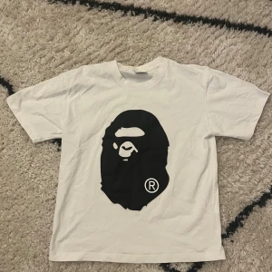 Vit BAPE t-shirt med ape-print - Vit t-shirt från BAPE med stort svart ape-ansikte tryckt på framsidan och en svart siluett på baksidan. Klassisk rund hals och korta ärmar. Tillverkad i Japan i mjuk bomull, perfekt för dig som gillar streetwear och vill sticka ut.