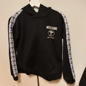 Svart Moschino hoodie med logoband - Svart hoodie orginal från Moschino Teen med vit logotyp på bröstet och vita band med Moschino-symbol längs ärmarna. Tröjan har huva, ficka vid magen och långärmad design. Snygg streetstil som sticker ut med coola detaljer. Den är köpt på rea för 1249 kr. 