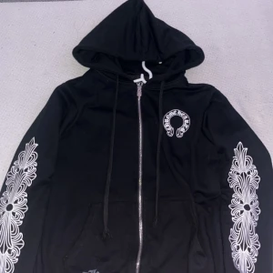 Svart Chrome Hearts hoodie med tryck - Svart hoodie från Chrome Hearts med vit tryckt logga på bröstet och detaljerade vita mönster längs båda ärmarna. Tröjan har huva, snörning och dragkedja framtill samt fickor. Materialet är mjukt och känns som bomull, perfekt för en chill och snygg streetwear-look.