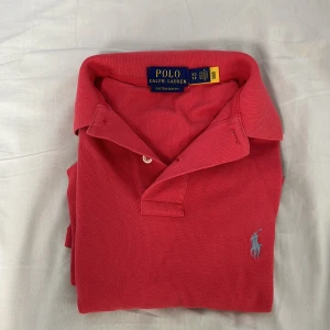 Röd pikétröja från Polo Ralph Lauren - Snygg röd pikétröja från Polo Ralph Lauren med klassisk krage och två knappar. Tröjan har korta ärmar och den ikoniska broderade loggan i ljusblått på bröstet. Modellen är custom slim fit och tillverkad i bomull för en skön känsla.