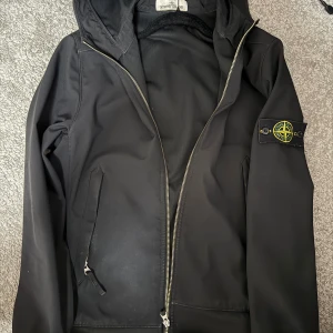 Svart softshelljacka Stone Island S - Svart softshelljacka från Stone Island med ikonisk patch på ärmen. Jackan har huva, dragkedja framtill och två fickor med dragkedja. Materialet är vatten- och vindtåligt polyester, perfekt för blåsiga dagar. Snygg och stilren design som funkar till streetwear.