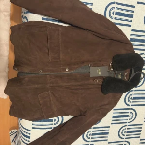 Brun mockajacka med päls från AGF Marostica - Äkta AGF Marostica Snygg brun mockajacka från AGF med stor svart pälsig krage och rejäla fickor framtill. Jackan har dragkedja och knappar, samt detaljer i metall vid kragen. Insidan är fodrad i grått tyg och har innerficka med dragkedja. Perfekt för dig som gillar klassisk stil med edge.