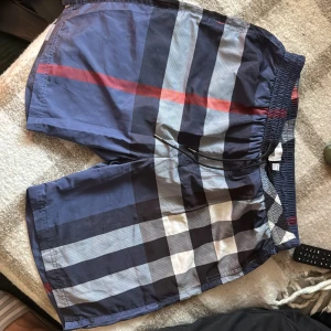 Rutig badshorts från Burberry XL - Snygga badshorts från Burberry i klassiskt rutigt mönster med blå, vit, svart och röd färg. Shortsen har elastisk midja med snörning och meshfoder. Perfekta för stranden eller poolen, med en stilren och lyxig känsla. Passar L också 