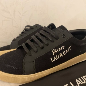 Svarta Saint Laurent sneakers - Snygga svarta sneakers från Saint Laurent med vit broderad logga på sidan. Skorna har svarta snören, detaljer i skinn och canvas samt en beige platt gummisula. Perfekt för dig som gillar stilrena och exklusiva sneakers med streetkänsla.