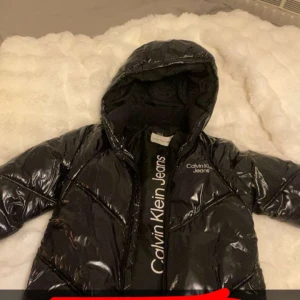 Svart pufferjacka från Calvin Klein Jeans - Säljer en svart, glansig pufferjacka från Calvin Klein med luva och vit logotyp på bröstet och insidan. Jackan har lång ärm och dragkedja framtill. Perfekt för dig som vill ha en snygg och varm jacka. Skriv till mig för mer bilder. 