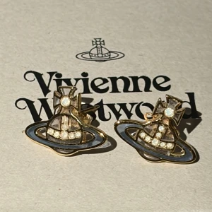 Äkta Vivienne Westwood örhängen guld - Ikoniska örhängen från Vivienne Westwood i guldtonad metall med glittriga stenar och den klassiska orb-designen i medium storlek med en unik och sällsynt design från deras sortiment. Små och stilrena, perfekta för att ge din look en edgy touch. Levereras med original ask och tygpåse med äktighets certifikat. Produkten är inköpt direkt från butik för 3000kr. Därav säljs för 1600kr