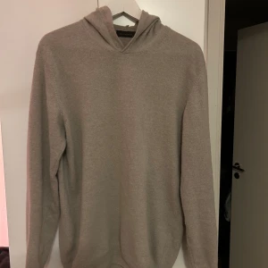 Beige hoodie från Oscar Jacobson M - Snygg beige hoodie från Oscar Jacobson i storlek M. Tröjan är tillverkad i Italien och har en mjuk, stickad känsla med huva och långa ärmar. Perfekt för dig som gillar stilrena och klassiska plagg med premiumkänsla. Använd ca 4 gånger